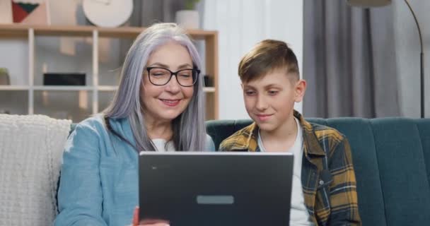 Довольно счастливая улыбающаяся добрая бабушка 65-летнего возраста обнимает своего милого радостного внука, когда они вместе смотрят интересную программу на компьютере дома — стоковое видео