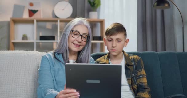 Agradable calma positiva de pelo gris abuelita jubilada y 12 años de edad niño relajado sentado con abrazos en el sofá y ver interesante espectáculo en el ordenador portátil — Vídeo de stock