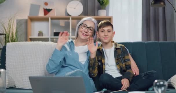 Nahaufnahme der gut aussehenden, fröhlichen, grauhaarigen Oma und des 12-jährigen Enkels, die zu Hause zusammen auf dem Sofa sitzen und Hände in die Kamera winken — Stockvideo