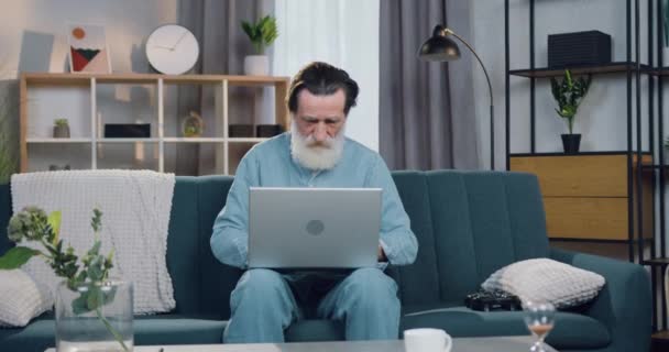 Atraktivní přemýšlivý chytrý starší vousatý muž sedící doma na pohodlné pohovce a pracující na počítači, pohled zepředu, zpomalený pohyb — Stock video