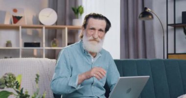 Evdeki rahat koltukta oturan ve kendine güveni tam bir yüz ifadesiyle kameraya bakan yakışıklı, pozitif sakallı yaşlı adamın ön görüntüsü.