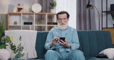 Çekici, pozitif, sakin, sakallı bir adamın portresi. Rahat evdeki yumuşak koltukta oturan ve telefon kullanarak arama yapan, ağır çekim.