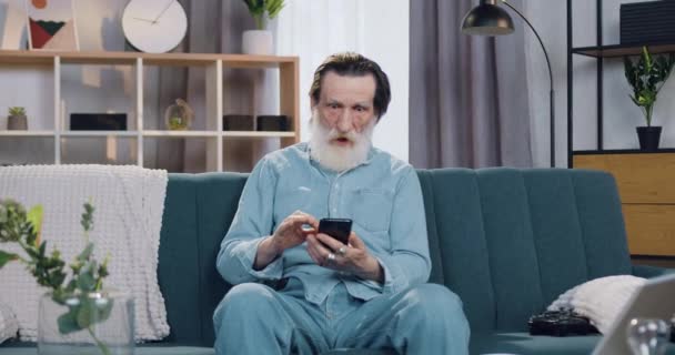 Attraktiv overrasket senior gråskægget mand fik gode nyheder på smartphone og glædeligt med løftede hænder sidder på sofaen derhjemme – Stock-video
