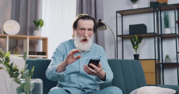 Surprised happy cheerful mature bearded man making big eyes when reading super news on his mobile and rejoycing from his winning in lottery at home — Stock Video