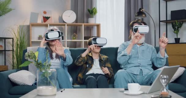 Retrato de atractivo abuelo moderno satisfecho y su nieto que se divierten utilizando auriculares de realidad virtual sentado en el sofá en el acogedor apartamento — Vídeos de Stock