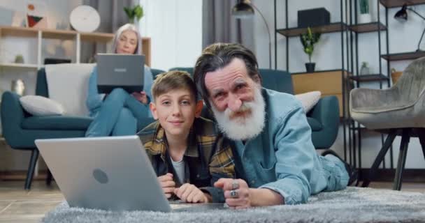 Guapo sonriente abuelo mayor barbudo feliz tumbado en el suelo junto con su nieto de 12 años de edad y el uso de la computadora portátil, posar en la cámara mientras la abuela sentado en el sofá y leer noticias en el ordenador — Vídeo de stock