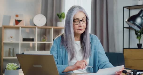 Чарівна спокійна впевнена у собі успішна 60-річна жінка, що сидить перед комп'ютером і перевіряє паперовий звіт з даними на екрані ноутбука, дистанційно концепція роботи — стокове відео
