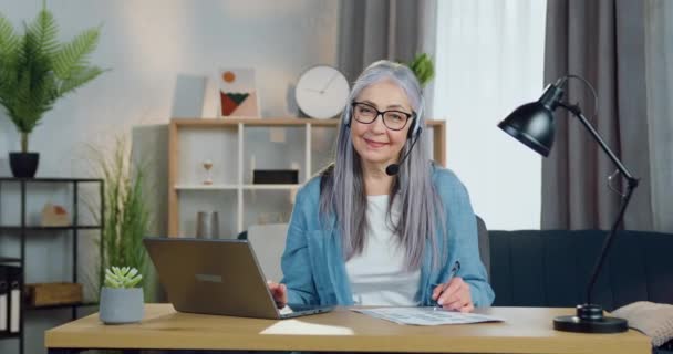 Linda sonriente feliz calificada mujer mayor de pelo gris en auriculares sentados delante de la cámara antes de la reunión de vídeo con compañeros de trabajo o clientes, concepto de trabajo distante — Vídeo de stock