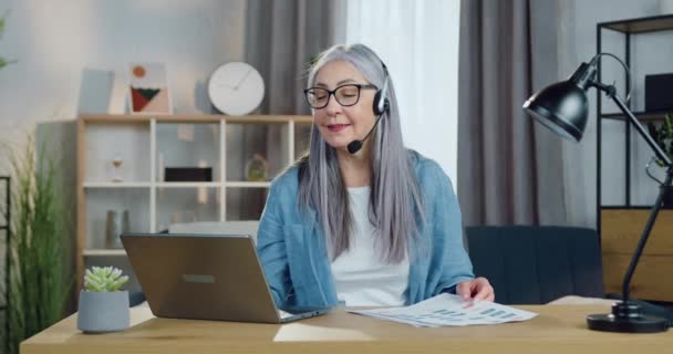 Conceito de trabalho à distância, onde adorável sorrindo mulher sênior confiante em fones de ouvido realizando reunião on-line no computador com colegas de trabalho do escritório em casa — Vídeo de Stock