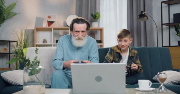 Attraktiv glad rolig glad glad skäggig senior farfar och hans 12-åriga sonson har kul tillsammans medan du spelar tv-spel på bärbar dator hemma — Stockvideo