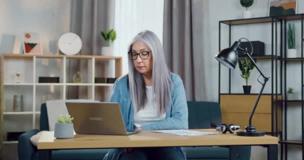 Attraente qualificata donna anziana dai capelli grigi positiva seduta sul suo posto di lavoro a casa e che lavora su laptop, lavoro a distanza dal concetto di home office — Video Stock