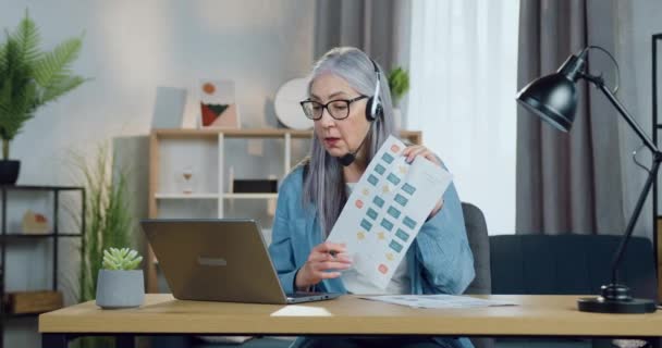 Lovely satisfied skilled mature woman in headset sitting at the table in home office during video conference and discussing report with clients or coworkers,remote work concept — Stock Video