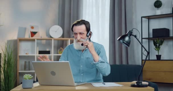 Portret van een goed uitziende emotioneel positieve bebaarde volwassen man in een headset die thuis achter de computer zit tijdens een videovergadering — Stockvideo