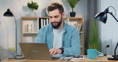 Çekici, kendine güvenen, sakallı bir genç adam evinde oturuyor ve bilgisayar ekranından kağıt raporuna notlar yazıyor.