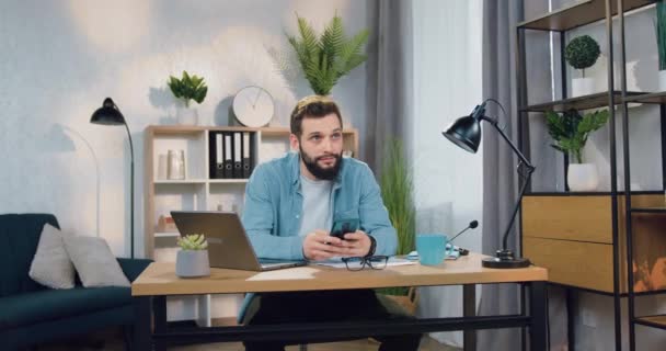 Dreamy handsome young bearded guy sitting at his workplace at home and uses his mobile to read news or write message,front view — Stock Video