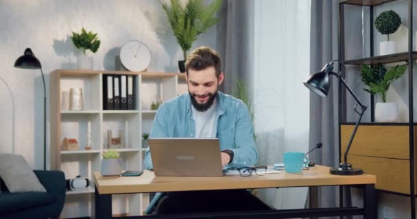 Красивый улыбающийся уверенный в себе опытный молодой бородатый мужчина в повседневной одежде работает на ноутбуке в красиво оформленном домашнем офисе — стоковое видео