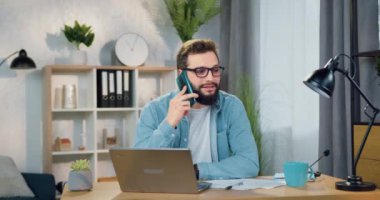 Yakışıklı, gülümseyen, gözlüklü, akıllı telefonlu genç adam güzel dekore edilmiş bir odada dizüstü bilgisayarla konuşurken konuşuyor.