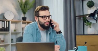 Yakışıklı, iyi modda, sakallı, gözlüklü, cep telefonuyla konuşan ev bilgisayarının önünde oturan, ağır çekimde konuşan.