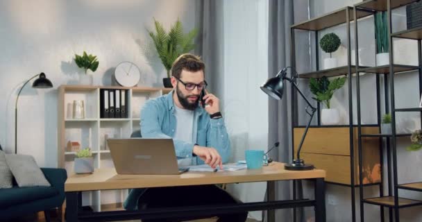 Atractivo confiado concentrado joven barbudo experto en gafas sentado en su lugar de trabajo en la oficina en casa y anotar datos mientras habla por teléfono — Vídeos de Stock