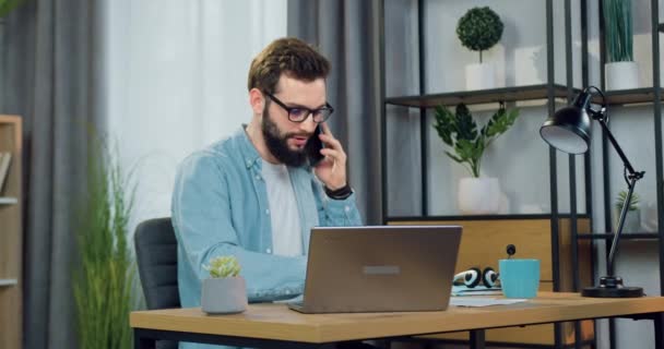 Sannolikt självsäker framgångsrik 30-årig skäggig manlig arbetstagare har mobil konversation när du sitter vid bordet på hemmakontoret och arbetar på datorn — Stockvideo