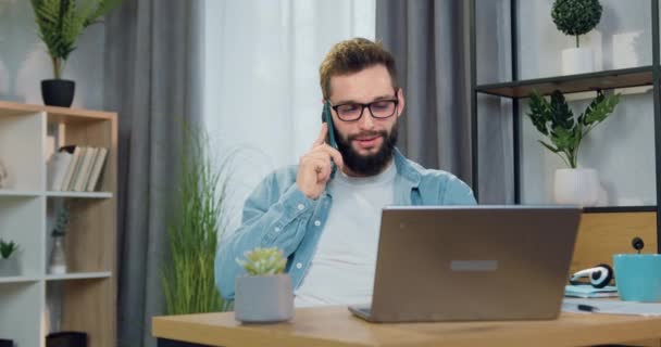 Közelkép a jóképű elégedett professzionális fiatal szakállas férfi munkavállaló szemüvegben, amely ül a számítógép előtt otthon, és beszél a mobil — Stock videók