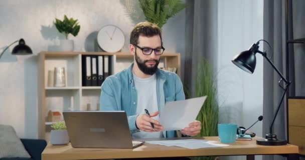 Retrato de bom aspecto confiante positivo experiente barbudo jovem em óculos que a verificação de relatórios de papel no local de trabalho no escritório em casa — Vídeo de Stock