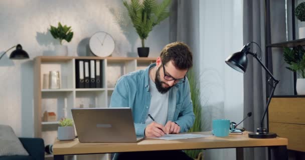 Красивый в хорошем настроении уверенный умный бородатый мужчина 30 лет сидит за столом в домашнем офисе и записывает важные заметки в бумажный отчет — стоковое видео