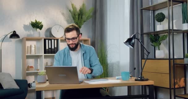 Bonito sorrindo jovem barbudo bem sucedido em óculos bebendo café ou chá enquanto trabalhava em laptop em casa, close-up, câmera lenta — Vídeo de Stock