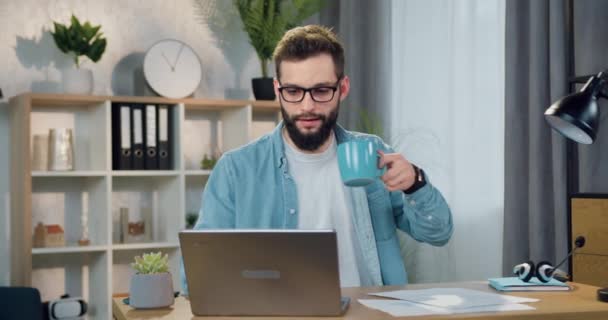 Портрет симпатичного уверенного опытного молодого бородатого парня в очках, который работает на ноутбуке дома и пьет чашку кофе — стоковое видео