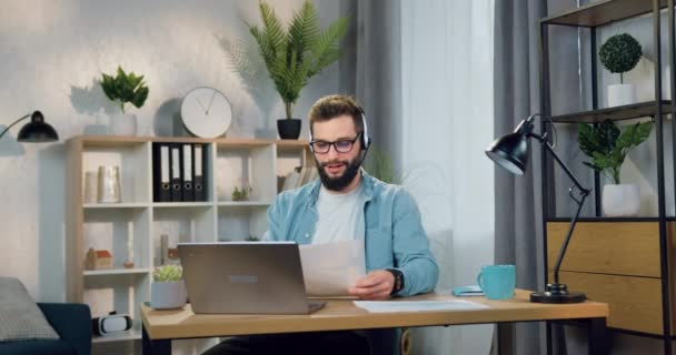 Conceito de negócio remoto onde atraente sorrindo bem sucedido jovem barbudo cara no fone de ouvido sentado na frente do computador durante o bate-papo por vídeo com colega de trabalho — Vídeo de Stock