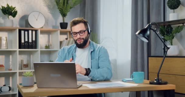 Привлекательный улыбающийся удовлетворенный успешный молодой бородатый парень в наушниках проводит видеочат с другом или коллегой, сидящим в домашнем офисе — стоковое видео