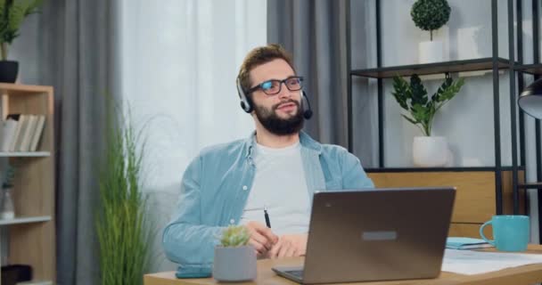 Симпатичный веселый расслабленный бородатый парень 30 лет в наушниках сидит перед компьютером во время видеозвонка с другом и наслаждается разговором — стоковое видео