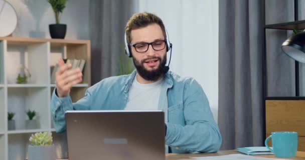 Bonito bem sucedido confiante jovem trabalhador barbudo no fone de ouvido sentado na frente do laptop durante reunião de vídeo com colega de trabalho ou cliente e discutir projeto conjunto — Vídeo de Stock