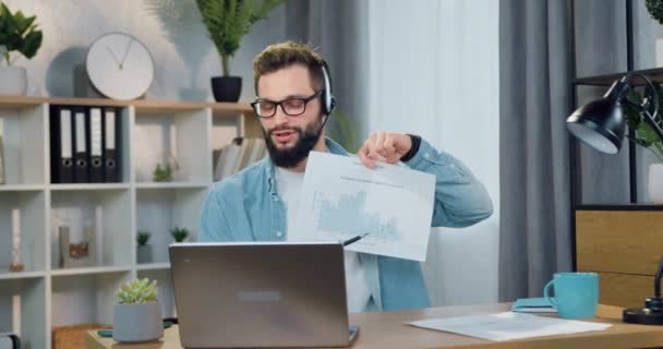 Atractivo positivo confiado joven barbudo capacitado en auriculares discutir informe con histograma durante la videoconferencia con compañeros de trabajo o clientes — Vídeos de Stock