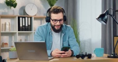 Çekici, gülümseyen, kulaklıklı, mutlu, sakallı genç bir adam. Evindeki iş yerinde çalan telefon listesinden müzik dinliyor.