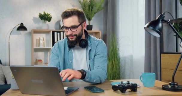 Улыбчивый довольный опытный 30-летний бородатый парень в наушниках, слушая музыку из плейлиста телефона и работая на компьютере из домашнего офиса — стоковое видео