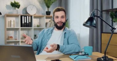 Güzel görünüşlü, sakallı, kulaklıklı, kameranın önünde oturan ve güzel dekore edilmiş oda arka planında video blogu kaydeden genç bir adam.