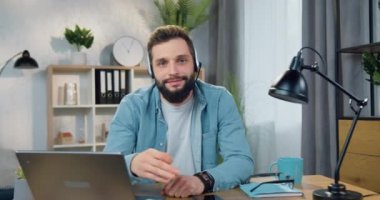 Güzel görünüşlü, gülümseyen, mutlu, sakallı, kulaklık takan, kameraya poz verip evinde oturmasını onaylayan genç adamın portresi.