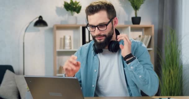 Όμορφος χαμογελαστός ανέμελος νεαρός γενειοφόρος με ακουστικά απολαμβάνοντας τη σύγχρονη μουσική, ενώ εργάζονται στον υπολογιστή στο γραφείο στο σπίτι, μπροστινή άποψη — Αρχείο Βίντεο