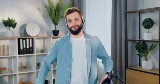 Портрет красивого улыбающегося уверенного молодого бородатого парня в наушниках, стоящего в красиво оформленной комнате и смотрящего в камеру со скрещенными руками — стоковое видео
