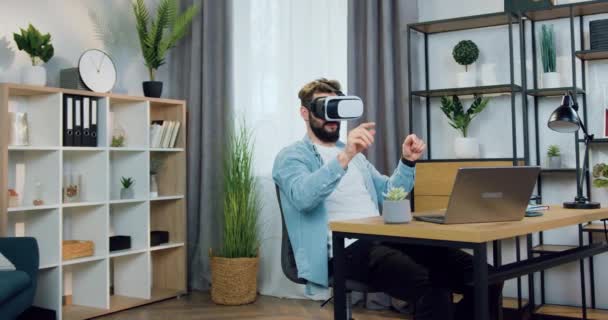 Симпатичный позитивный умный опытный молодой бородатый человек сидит в домашнем офисе и работает в гарнитуре дополненной реальности, современная концепция технологии — стоковое видео