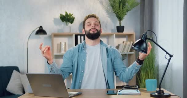 Retrato de simpático jovem barbudo relaxado que sentado em seu local de trabalho em casa e meditando com os olhos fechados para desfrutar de harmonia e silêncio em torno dele — Vídeo de Stock