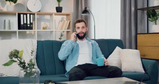 Portret van knappe in goed humeur kalme jonge berded man die zitten op de bank thuis en praten op de smartphone gelijktijdig het drinken van koffie — Stockvideo