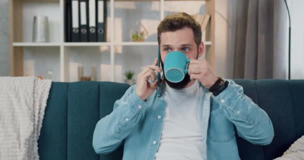 Aangename glimlachende vrolijke jonge bebaarde persoon die thuis thee of koffie drinkt en praat op smartphone, close-up — Stockvideo