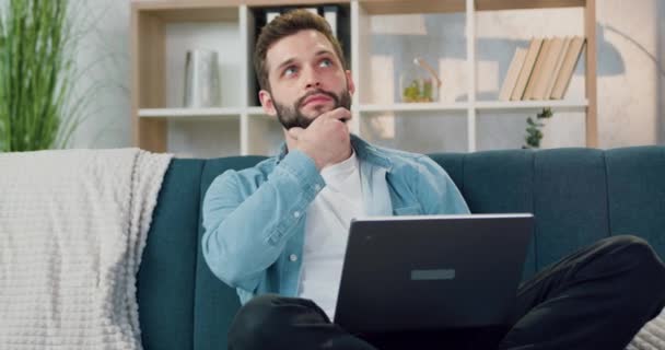 Soñador guapo despreocupado joven barbudo sentado en un sofá suave en casa y trabajando en el ordenador, vista frontal — Vídeos de Stock