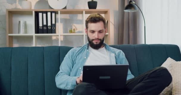 Привлекательный улыбающийся опытный бородатый парень сидит в расслабленной позе на мягком диване дома и доволен результатами проделанной работы на компьютере — стоковое видео
