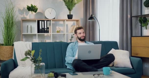 Provável sonhador despreocupado beared cara em roupas casuais sentado em pose relaxada no sofá confortável no quarto aconchegante e trabalhando no laptop, close up — Vídeo de Stock