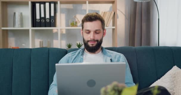 Vista frontale di bello soddisfatto sorridente giovane uomo barbuto che si rilassa con mettere le mani dietro la testa dopo aver finito il suo lavoro sul computer portatile a casa — Video Stock