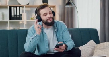 Çekici, rahat, gülümseyen, kulaklıklı, sakallı, modern melodileri dinleyen, evdeki yumuşak koltukta poz veren genç adam.