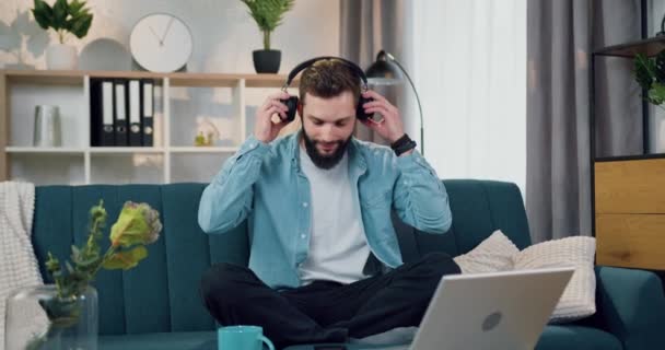 Agradable sonriente despreocupado joven barbudo sentado en cómodo sofá en casa y disfrutando de la música de la lista de reproducción del teléfono con auriculares — Vídeos de Stock